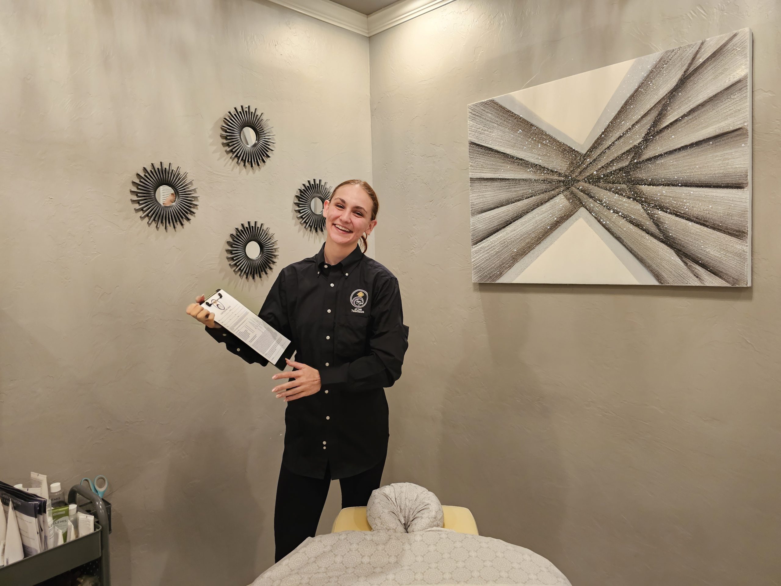 A smiling person in a black uniform holds a clipboard in a room decorated with abstract wall art and mirrors. A covered massage table is in front of them, ready for the next relaxing spa treatment.