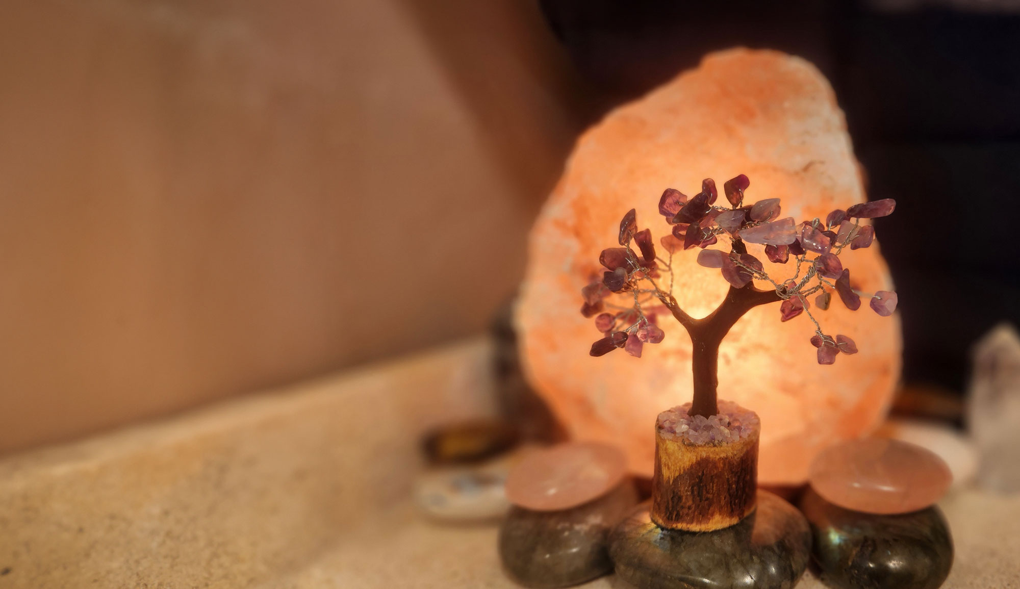 A small decorative tree with purple gemstones is placed in front of a glowing Himalayan salt lamp on a beige surface, creating a serene spa-like ambiance perfect for relaxation, massage, or facials.