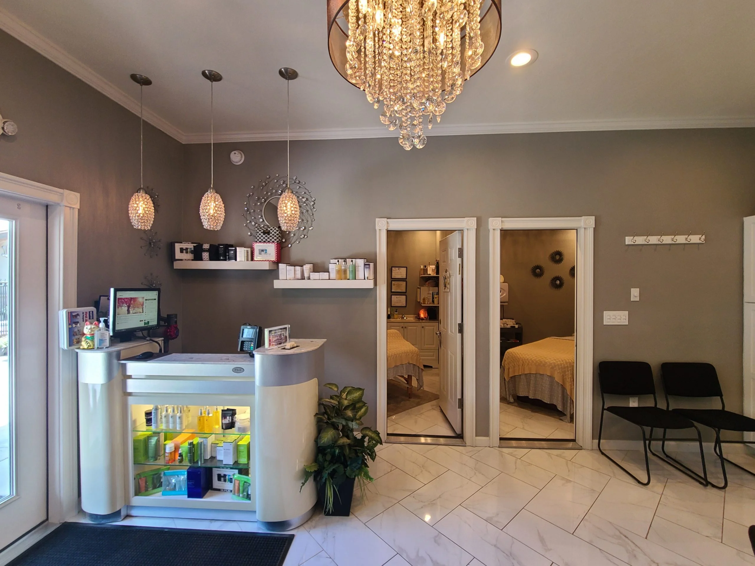 The well-lit interior of the spa features a reception counter with beauty products, a chandelier, three pendant lights, two treatment rooms for massages or facials, and a small seating area with two black chairs.