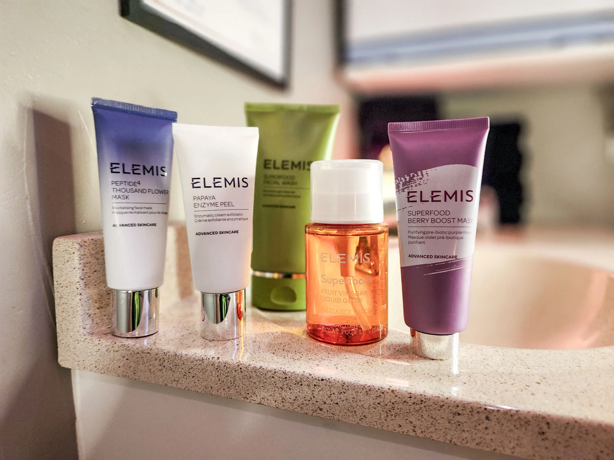 Five Elemis skincare products arranged on a bathroom counter, including tubes and a small orange bottle.