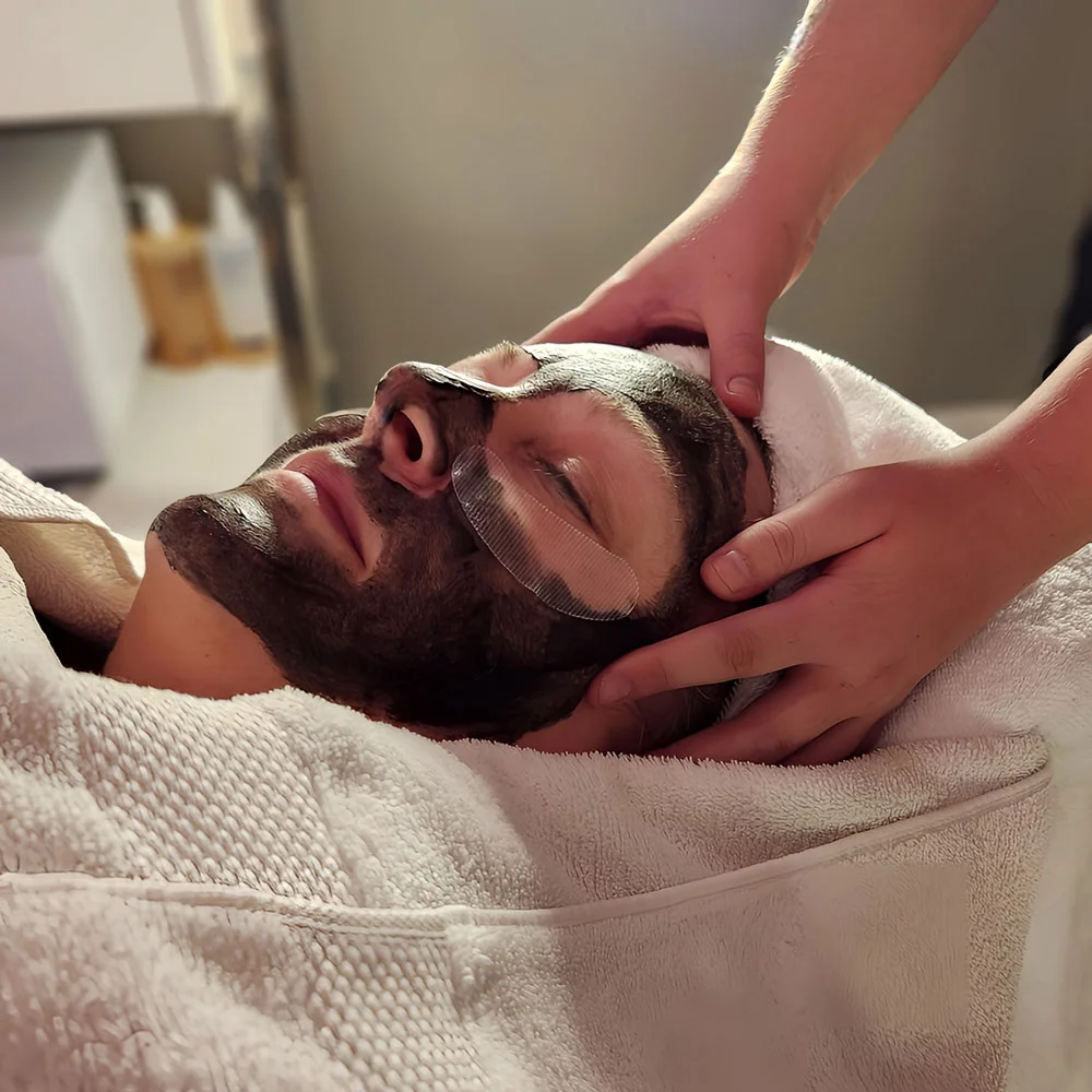 Person receiving a facial treatment with a dark face mask and eye patches while lying under a towel.